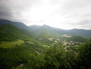 Obraz premium Panorama montano con alberi in primo piano, valle verde e piccolo centro abitato sullo sfondo, incorniciato da colline e montagne sotto un cielo nuvoloso; atmosfera naturale e tranquilla