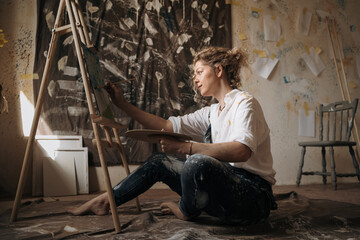 Fototapeta premium Education, creation, art and talent. Young curly painter woman wearing jeans overalls painting on canvas with paints creative hobby sitting on floor in creativity workshop.