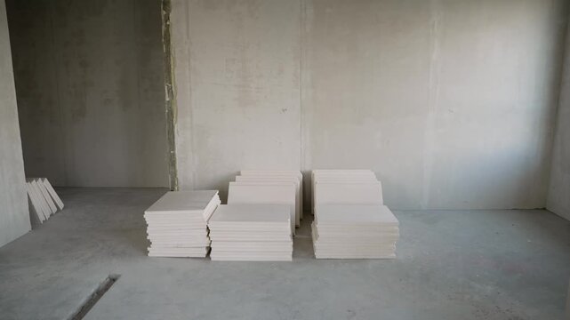 Empty room under construction shows renovation process. Raw concrete walls floor with stacked white plasterboard. Natural light from side window creates minimalist, industrial background