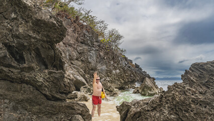 A man stands in the surf on a tropical beach, between rocks, smiling. The hand is raised up. The waves of the turquoise ocean are foaming on the sand. Clouds in the sky. Philippines. Palawan island. © Вера 