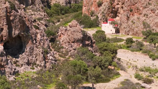 Agiofarago Gorge Crete Greece 70mm close up aerial of massive cave in orange limestone cliff, drone flight towards mountain cavern and remote chapel, rocky texture and geological formations