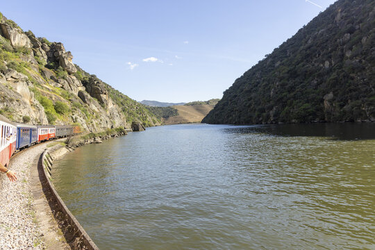 View of the Douro River with a train traveling along its banks through steep rocky hills and lush vegetation under a clear blue sky in Portugal.
