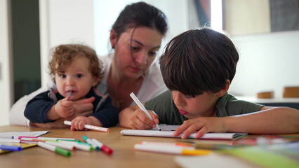 Fototapeta premium Mother with toddler and older child drawing together at table during calm creative moment as boy concentrates on artwork while baby explores markers in warm-family setting