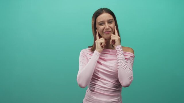Young hispanic woman pressing index fingers to cheeks while puckering then smiling in a teal studio backdrop; playful expression.