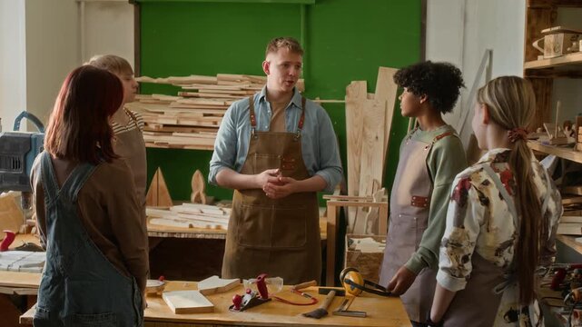 Young teacher giving instructions to teenage students listening to him carefully while all standing around table in woodworking workshop