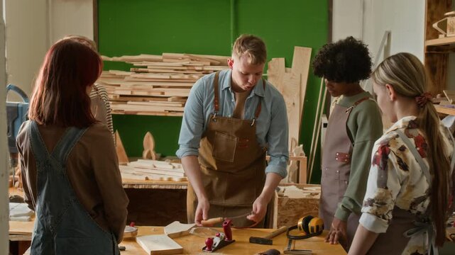 Young craftsman instructing group of teenagers on safety rules and showing coping saw to them in carpentry workshop