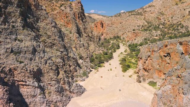 Agiofarago Gorge Crete Greece aerial pull back flight from canyon to turquoise beach, orange cliffs and hikers on sandy path, cinematic reveal of Mediterranean sea and mountains, travel discovery
