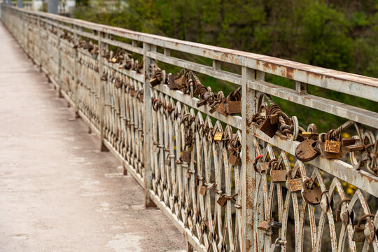 View of numerous rusty love locks attached to a metal bridge railing with lush green trees in the background in Kamianets-Podilskyi, Khmelnytska oblast, Ukraine.
