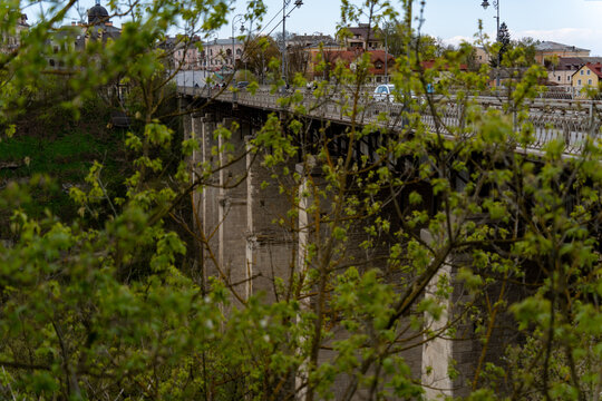 View of the Novoplanivskyi Bridge framed by green spring foliage with the historic old town buildings in the background in Kamianets-Podilskyi, Khmelnytska oblast, Ukraine.