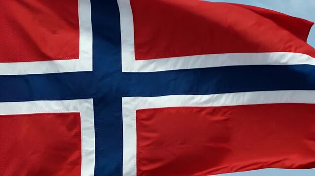 A close-up shot shows the flag of Norway in motion, showcasing its iconic design. The red, white, and blue cross pattern is clearly visible against a bright blue sky.