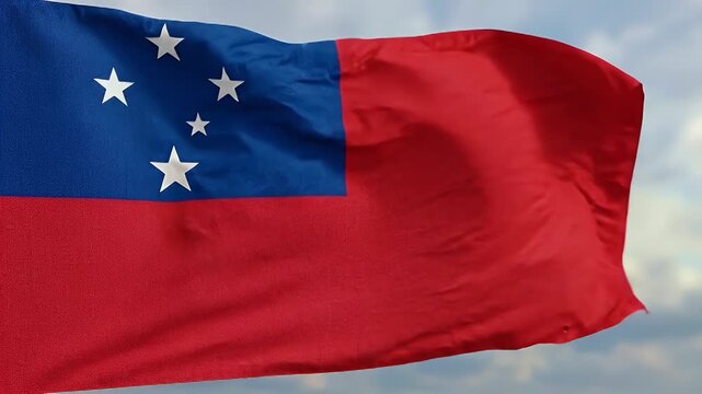 The national flag of Samoa is shown gracefully waving against a soft, cloudy sky. The flag displays a blue rectangle in the top corner with white stars and a larger red field, symbolizing the
