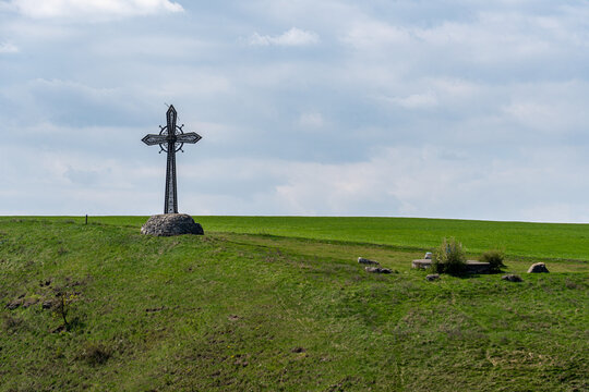 View of an ornate metal cross standing on a stone base atop a grassy hill under a vast cloudy sky in Kamianets-podilskyi, Khmelnytska oblast, Ukraine.