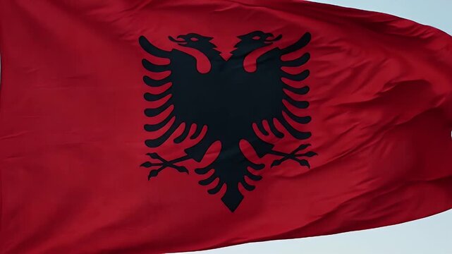 A vibrant red flag of Albania is seen waving in the wind. The flag features a bold black double-headed eagle at its center.