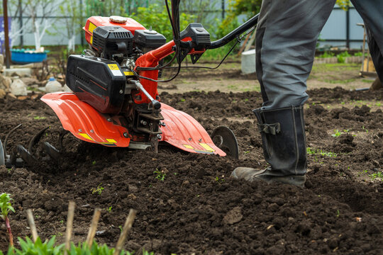 Farmer in rubber boots tilling garden soil with gasoline cultivator. A close-up action shot showing a person's leg in a protective rubber boot while operating a red power tiller. 