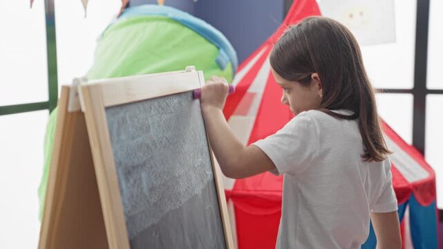 Girl erases chalkboard with eraser at easel in a children's playroom inside a bright building; quiet concentration.