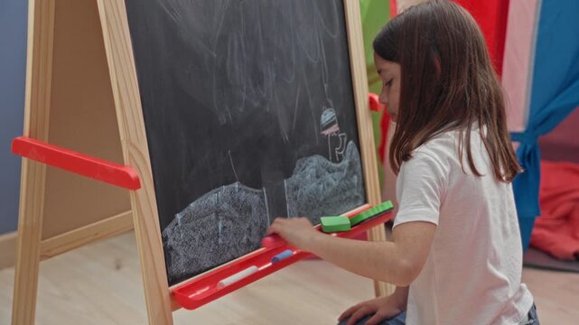 Girl draws on a freestanding chalkboard with hand and chalk beside eraser and wooden easel in classroom play area; concentration learning.