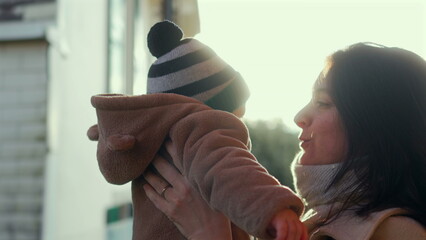 Mother embracing baby in bear teddy fleece coat and striped beanie hat outdoors, woman in beige coat and scarf holding child with eyes closed in moment of family tenderness © Marco