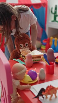 Girl placing a stuffed monkey and toy sandwich on a red play table amid dolls and plastic dishes in building; playful imagination.