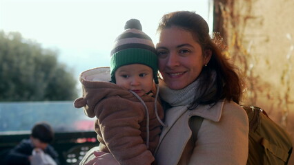 Mother holding baby in bear teddy fleece coat and striped beanie hat during winter day outdoors, family enjoying seasonal travel and togetherness with warm and happy expressions © Marco