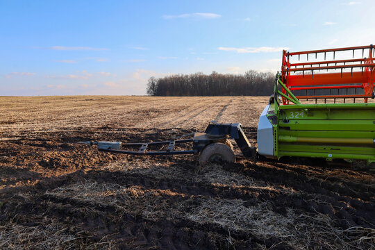 An agricultural trailer with a combine harvester header is stuck deep in wet soil in the middle of a mown field under a clear sky. Heavy machinery is bogged down in mud, demonstrating challenges durin