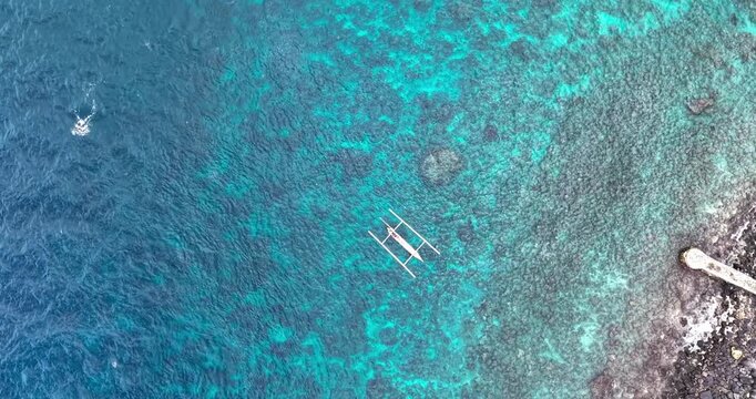 Aerial top-down drone shot of turquoise coral reef waters along the rocky coastline of Amed, Bali, Indonesia with a traditional Balinese fishing boat floating offshore.