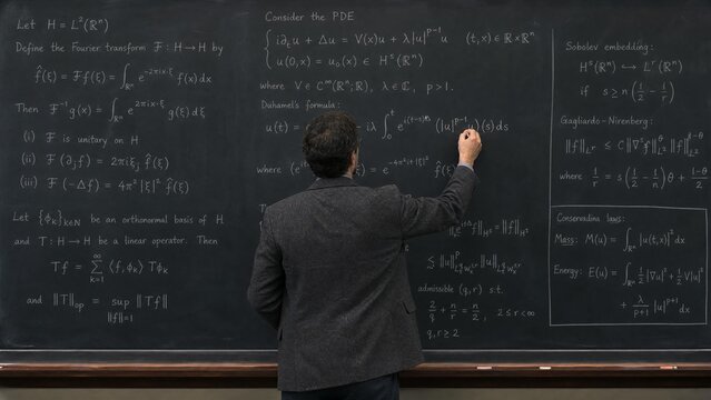 Male Professor Writing Complex Mathematical Physics Formulas on Blackboard in Classroom