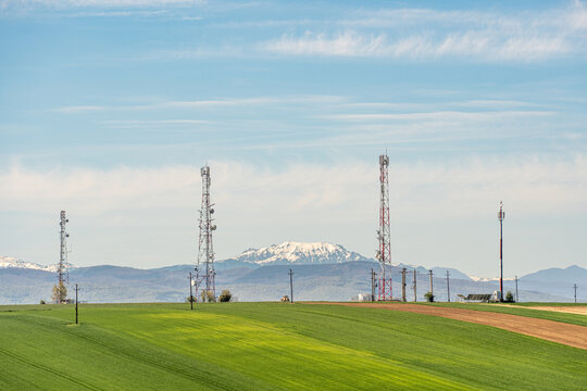 View of telecommunication towers standing in green fields with snow-capped mountains under a blue sky.