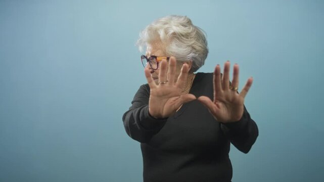 Senior woman wearing glasses and rings holds hands palms out in a stop gesture in studio blue backdrop; defensive refusal.