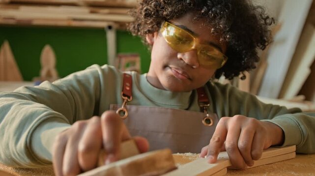 Rack focus view of passionate curly-haired African American teenage boy with yellow protective goggles sanding wooden piece in carpentry workshop