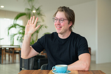 Authentic portrait of a happy Scandinavian man waving hi while sitting in a cafe with glasses. © Ranta Images