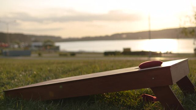 Red bean textile bag flies, slides across wooden cornhole board and drops into hole at sunset. Scenic shot of cornhole equipment on hill in park near lake during game at sunset.