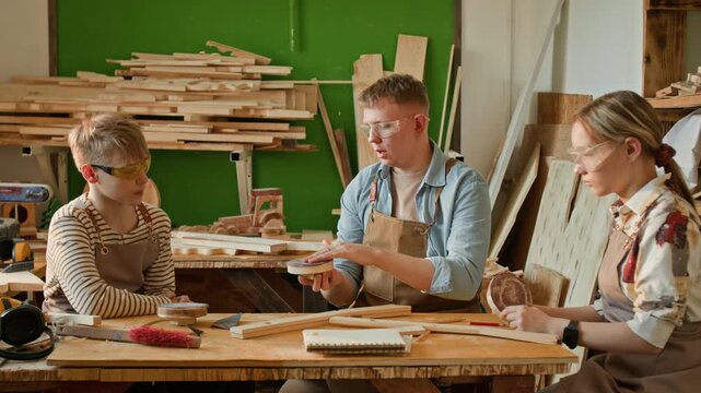 Gen Z woodworking instructor teaching two teenagers to polish wood with hand sander while all sitting at workbench during craft lesson