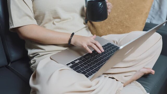 Woman casually dressed, sitting crossed legged on a sofa, remote working on her laptop and typing, holding a coffee mug and embracing a comfortable work life balance from home
