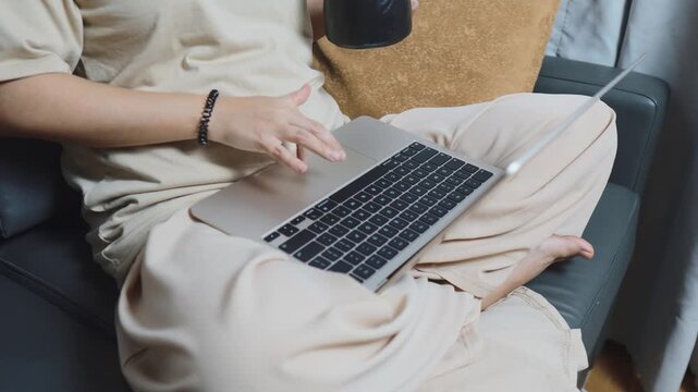 Person working remotely on a laptop while comfortably sitting on a couch in a casual setting, multitasking with a drink, demonstrating a flexible and connected work from home lifestyle