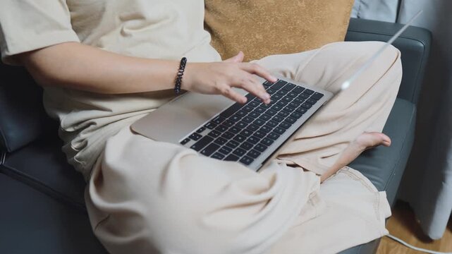 Person working from home, casually dressed, sitting on a dark grey sofa with a laptop on their lap, typing on keyboard and using technology for remote work, enjoying a cozy and flexible lifestyle