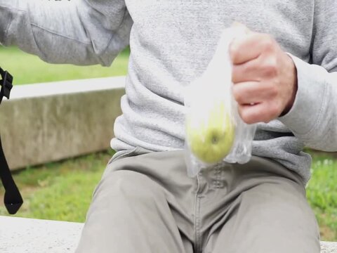 A close-up of a man sitting outdoors, removing a fresh apple from a thin plastic bag, highlighting the prevalence of plastic packaging in food.