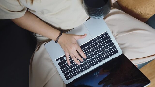 Woman working remotely on a laptop, hands typing on keyboard, holding a coffee cup, managing business tasks from a comfortable home office setup, focusing on productivity and freelancing flexibility