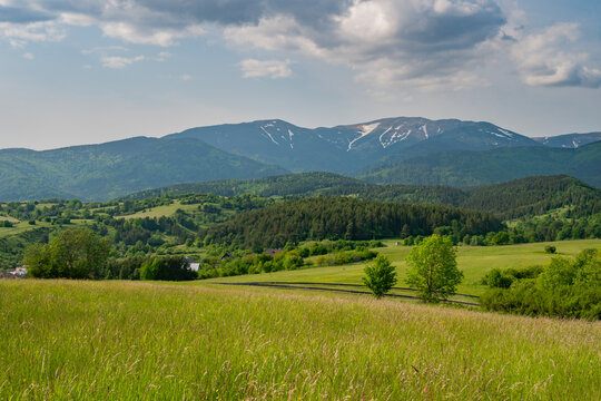 View of lush green meadows and rolling hills with the snow-capped peaks of the Low Tatras mountains under a cloudy sky in Horna Lehota, Banskobystricky kraj, Slovakia.