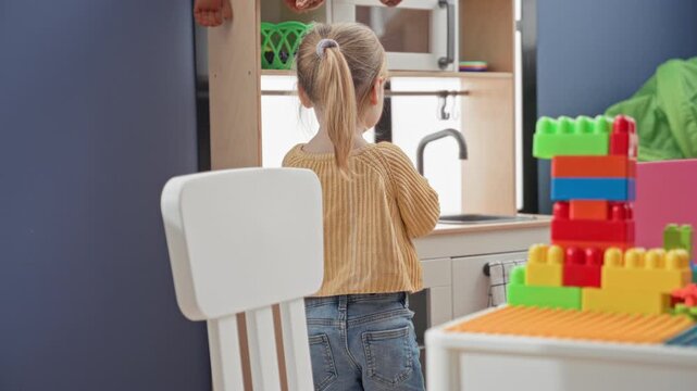 Girl with ponytail playing with colorful blocks beside a toy kitchen sink in building wearing yellow sweater and jeans; curiosity imagination.
