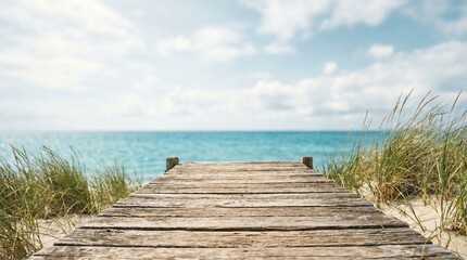 Obraz premium Wooden walkway leads through dune grass toward a quiet blue sea beneath a bright sky, creating a simple coastal access scene with open space and a peaceful summer atmosphere.