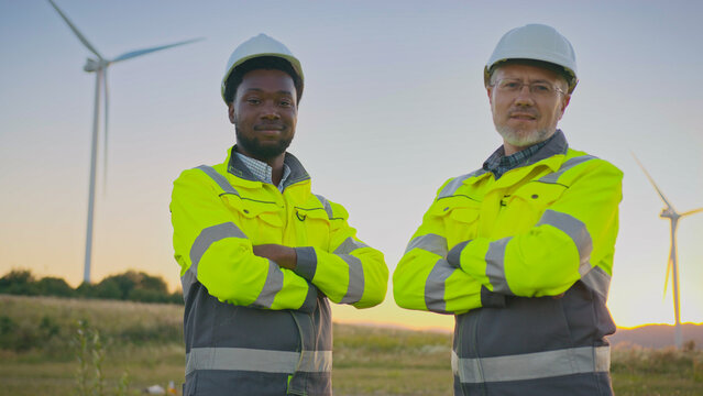 Two male workers in safety helmets and fluorescent jackets standing at wind turbines. Both crossing arms while facing camera. Professionals overseeing sustainable energy station in open field.
