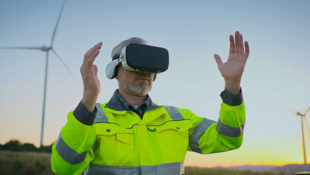 Male engineer in reflective jacket wearing VR headset at wind farm. Man raising hand and operating with gesture control. Worker using virtual reality technology for renewable energy project.