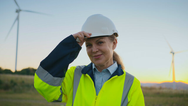 Confident female engineer wearing reflective jacket standing near wind turbines in open field. Woman touching hair and smiling at camera during sunset. Worker posing at renewable energy site.
