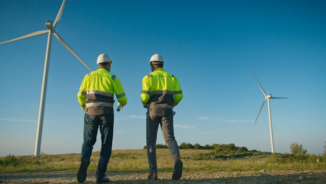 Two workers in helmets and reflective jackets standing in open field near wind turbines. Holding rolled documents and gesturing while talking. Colleagues discussing renewable energy project outdoors.
