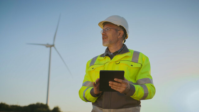 Male engineer in helmet and reflective jacket holding tablet. Worker standing outdoors in field near wind turbine. Man reviewing renewable energy project data and analyzing technical details.