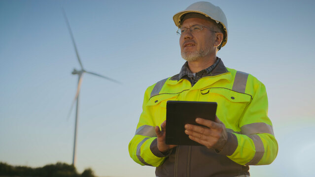 Worker standing outdoors in field near wind turbine. Man reviewing renewable energy project data and analyzing technical details.
