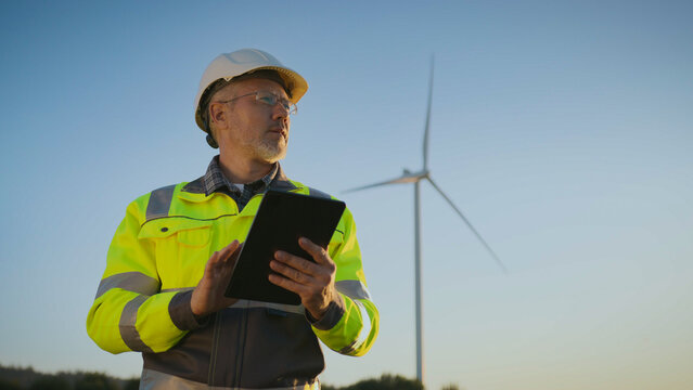 Male inspector in protective helmet and safety jacket holding tablet. Worker examining renewable energy documentation near wind turbine. Man analyzing wind farm project outdoors in field.