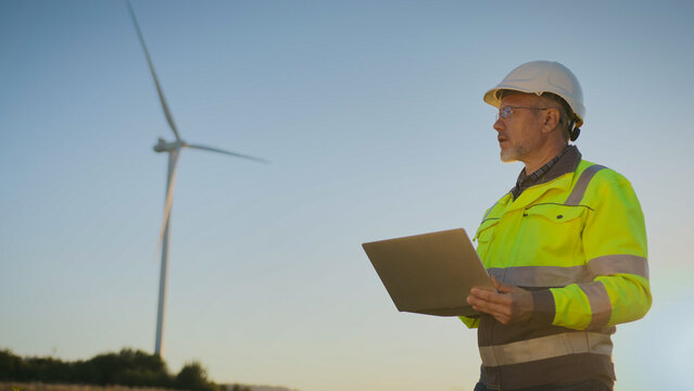 Male worker in helmet and reflective jacket holding laptop. Engineer standing in field near wind turbine. Monitoring renewable energy system. Man observing wind farm operations outdoors.