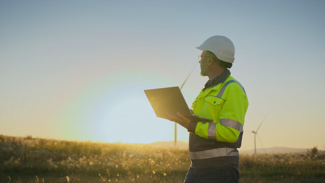 Male engineer in helmet and reflective jacket holding laptop in hands. Worker standing in field with wind turbine at sunset. Looking to side. Technician monitoring renewable energy project.
