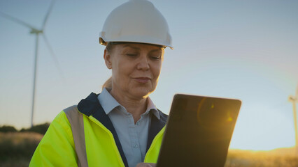 Fototapeta na wymiar Female worker in safety helmet and reflective jacket standing outside holding tablet. Woman reviewing wind energy project. Engineer checking data near turbine in countryside field at sunset.